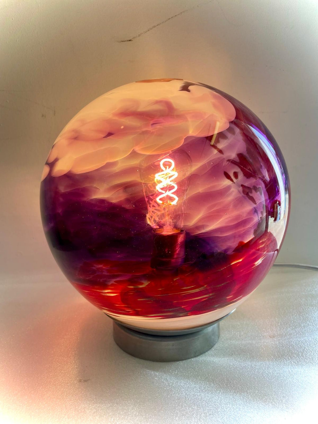A spherical table lamp featuring vivid swirling hues of pink, purple, and red glass, with a glowing filament bulb at its core, set on a metallic base and casting a warm, atmospheric glow.