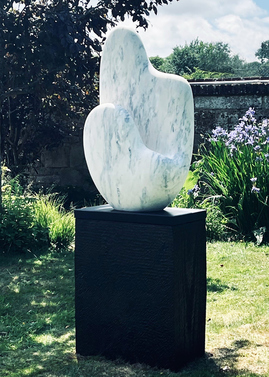 Jason Mulligan sculpture titled KINDRED, a one-off contemporary abstract form carved from Crema Aurora marble. The sculpture features a tall, dual-peaked organic shape with a softly curving hollow, evoking themes of kinship, connection, and balance. The marble surface is smooth and polished, with pale cream tones subtly veined with soft grey and warm blush accents, enhancing its natural elegance. Mounted on a tall dark plinth, the work is designed for both outdoor garden placement and indoor display.