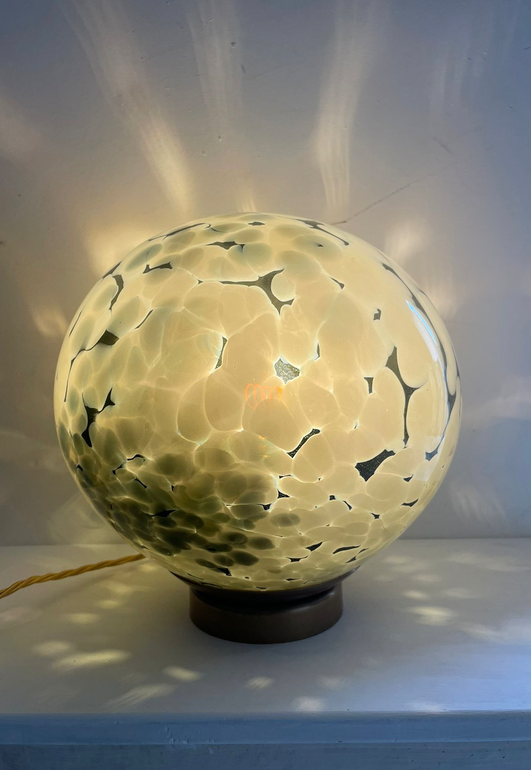 A spherical artisan table lamp with a mottled cream and dark green glass design, softly glowing and casting intricate patterns of light onto the surrounding surfaces.