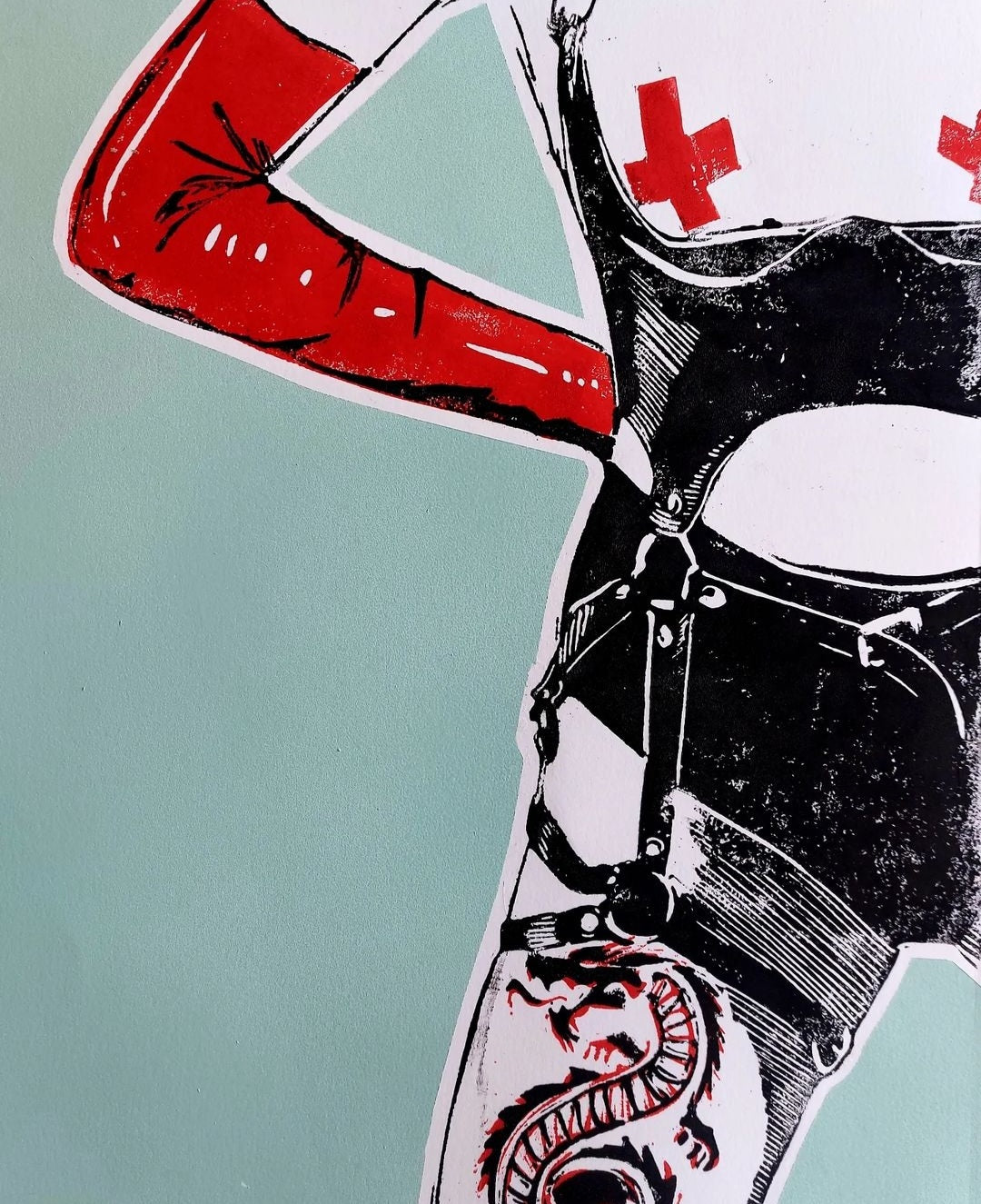 Cropped linocut artwork of a woman in bondage-style lingerie with bold red cross pasties, red opera gloves, and a red dragon tattoo spiralling down her thigh. The figure is rendered in black and white with sharp detailing, set against a cool mint green background. A visually striking, fetish-inspired composition blending tattoo culture with graphic sensuality.