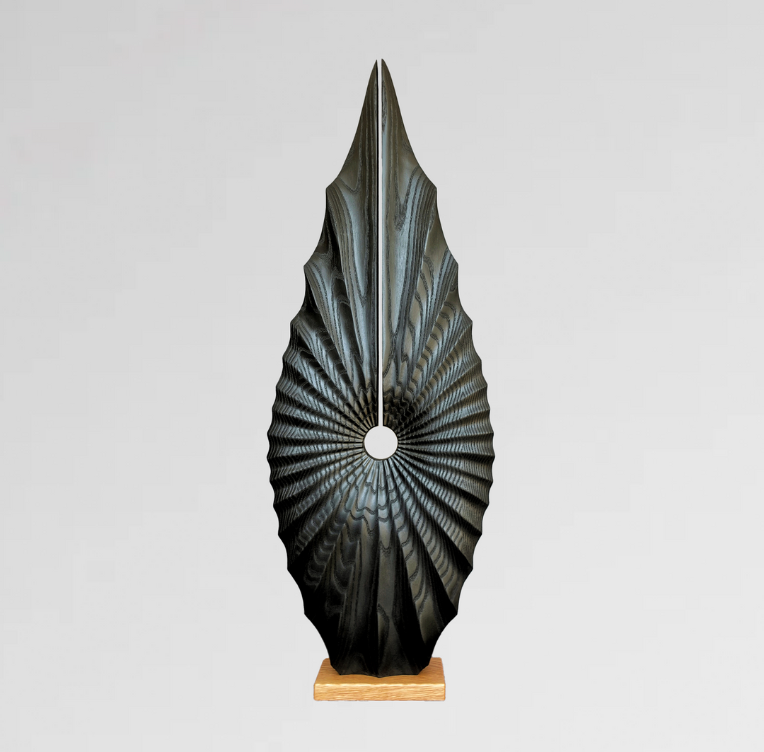 Tall abstract wood sculpture with radiating carved pattern, dark finish, and central circular void on a light wood base.