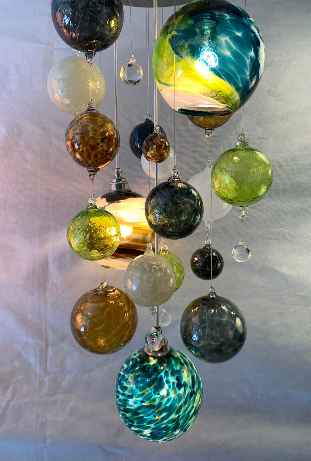 Artisan glass chandelier composed of suspended spherical orbs in rich tones of teal, green, amber, and white, with softly diffused internal lighting creating reflections across a neutral backdrop.