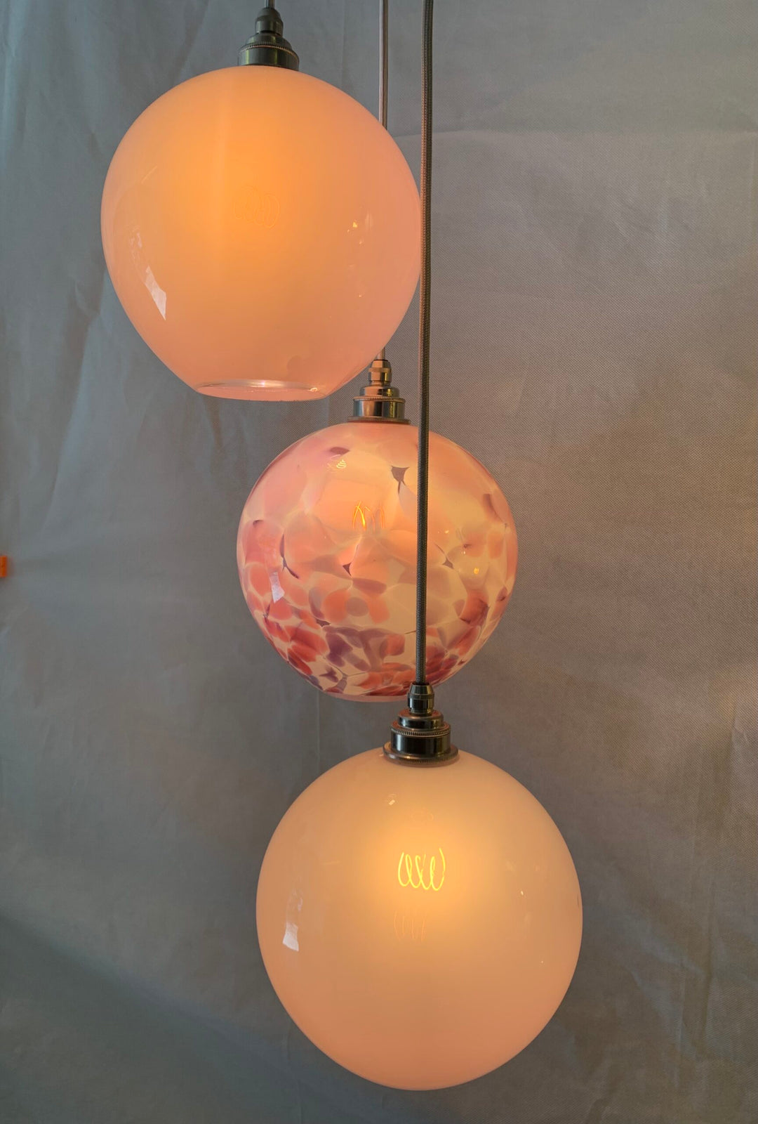Trio of artisan pendant lights with two opaque blush glass globes and one centrepiece globe featuring a delicate pink and rose-marbled pattern, suspended against a soft grey backdrop.