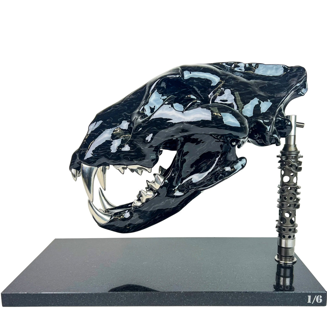 A striking, polished carbon fibre lion skull sculpture by Alastair Gibson, crafted with Mokume-style carbon layup for a swirling, chaotic texture. The jaw is fitted with gleaming bismuth alloy teeth. Mounted on a genuine F1 gearbox selector barrel atop a granite base, the work merges natural form with elite motorsport engineering.