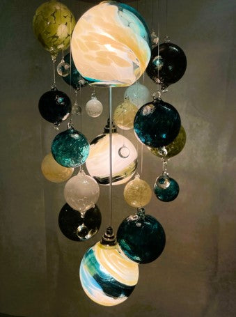 A sculptural glass chandelier composed of suspended hand-blown glass spheres in teal, olive, white, and smoky black tones, with two illuminated globes featuring marbled swirls of gold and aqua.
