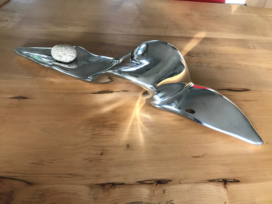 Sylph by Penny Hardy — a limited edition abstract sculpture with flowing, aerodynamic lines in polished metal, suggestive of movement and grace, accompanied by a smooth natural stone and displayed on a wooden surface.