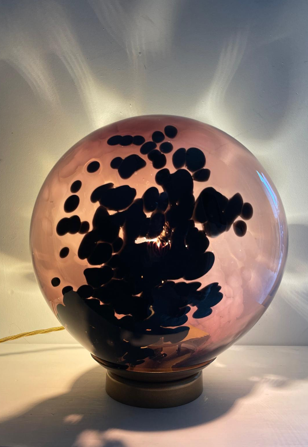 A handcrafted spherical table lamp made of pink glass with inky black organic patterns swirling within, set on a minimalist brass-toned base and casting soft light and shadows against the wall.