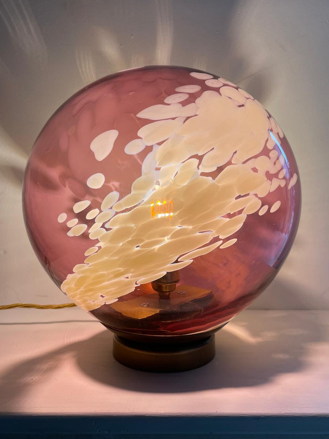 A soft pink glass table lamp with creamy white swirling patterns reminiscent of clouds, set on a bronze-toned base, radiating warm ambient light and delicate reflections on the wall.