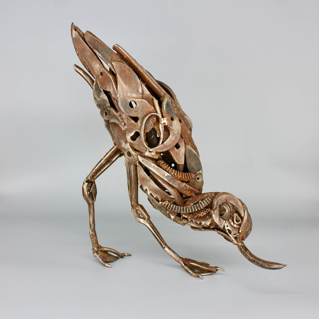 A dynamic sculpture of a bird pecking the ground, masterfully constructed by Mike Thompson using vintage tools and reclaimed metal. The figure is intricately assembled from spanners, gears, pliers, and other mechanical parts, forming a lifelike pose with a powerful, expressive stance. The surface bears a rich patina, enhancing the aged, industrial feel while celebrating the artistry in upcycling.