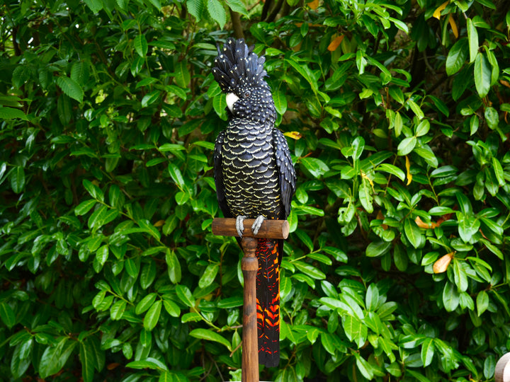 A highly detailed paper sculpture of a Red-tailed Black Cockatoo perched confidently on a wooden post, with jet black plumage dotted in pale yellow and ivory, a curved bone-coloured beak, and bold red-orange tail feathers, set against lush green foliage.
