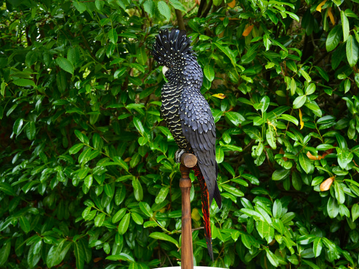 A highly detailed paper sculpture of a Red-tailed Black Cockatoo perched confidently on a wooden post, with jet black plumage dotted in pale yellow and ivory, a curved bone-coloured beak, and bold red-orange tail feathers, set against lush green foliage.