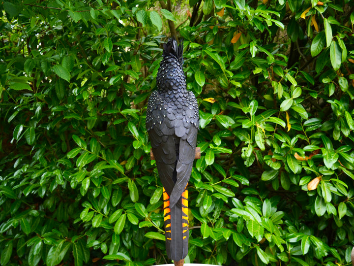 A highly detailed paper sculpture of a Red-tailed Black Cockatoo perched confidently on a wooden post, with jet black plumage dotted in pale yellow and ivory, a curved bone-coloured beak, and bold red-orange tail feathers, set against lush green foliage.