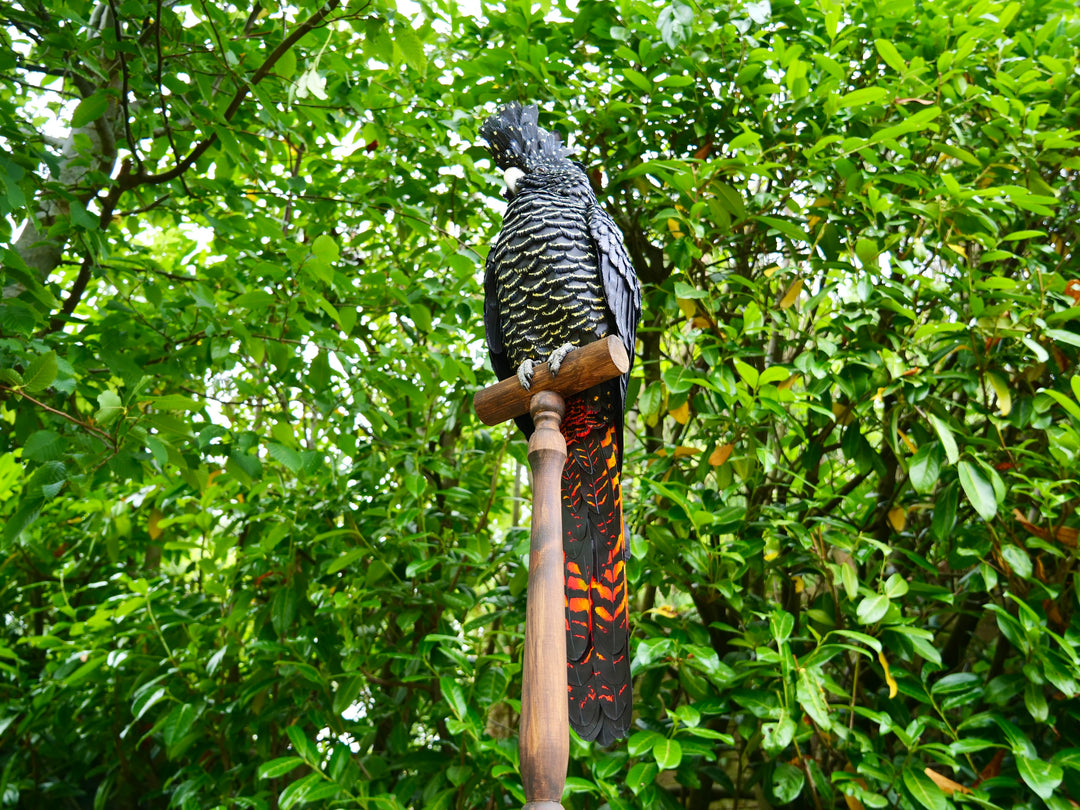 A highly detailed paper sculpture of a Red-tailed Black Cockatoo perched confidently on a wooden post, with jet black plumage dotted in pale yellow and ivory, a curved bone-coloured beak, and bold red-orange tail feathers, set against lush green foliage.