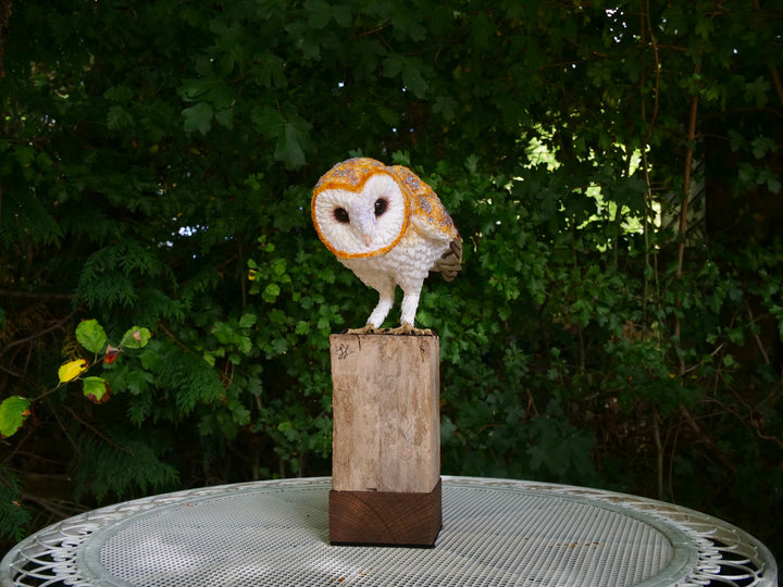 Hyperrealistic sculpture of a barn owl, perched on an oak block with its head gently tilted, featuring intricately layered ivory-white and golden-orange paper feathers with speckled grey detailing, set against a natural woodland backdrop.