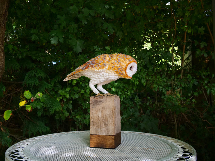 Hyperrealistic sculpture of a barn owl, perched on an oak block with its head gently tilted, featuring intricately layered ivory-white and golden-orange paper feathers with speckled grey detailing, set against a natural woodland backdrop.