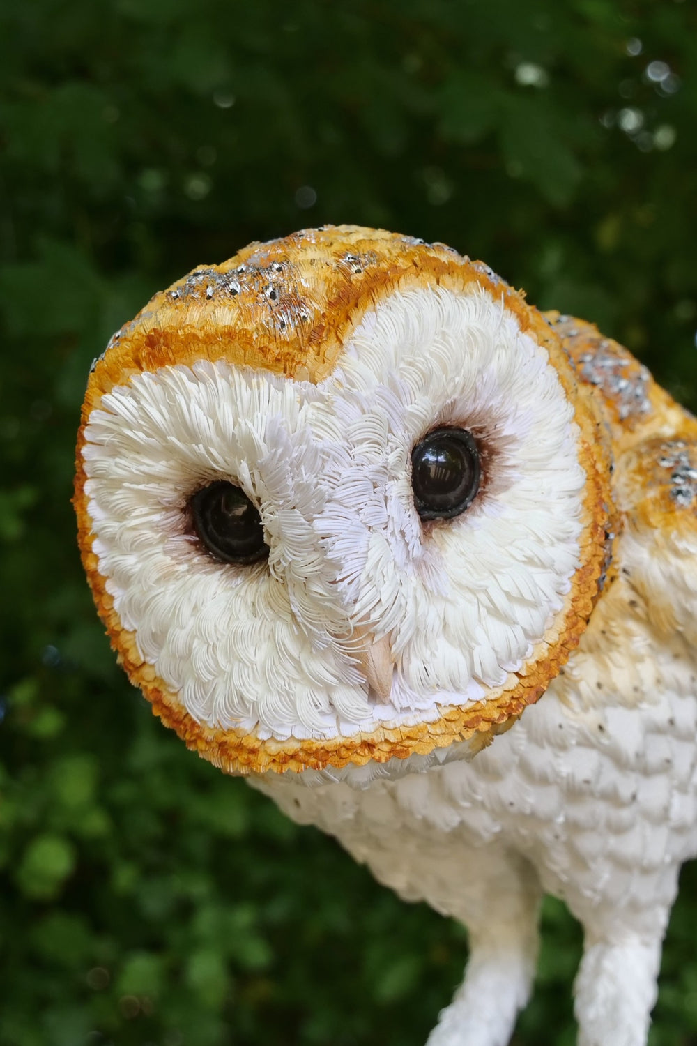 Hyperrealistic sculpture of a barn owl, perched on an oak block with its head gently tilted, featuring intricately layered ivory-white and golden-orange paper feathers with speckled grey detailing, set against a natural woodland backdrop.