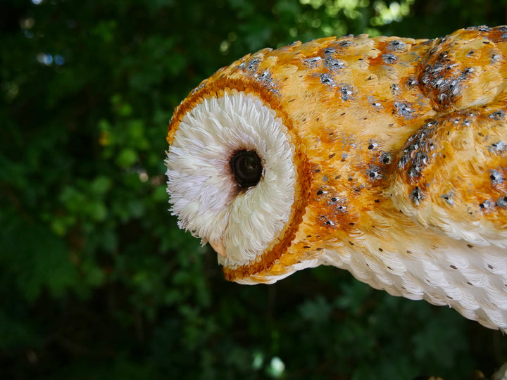 Hyperrealistic sculpture of a barn owl, perched on an oak block with its head gently tilted, featuring intricately layered ivory-white and golden-orange paper feathers with speckled grey detailing, set against a natural woodland backdrop.