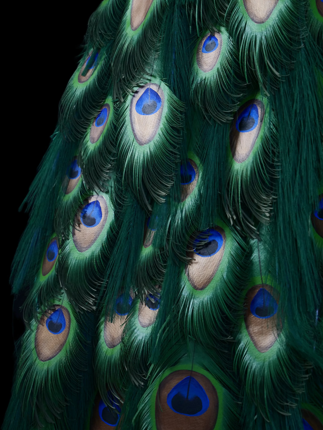 Arrogant Beauty by Jamie Lord – a life-size peacock sculpture meticulously crafted from hand-cut, hand-painted 260gsm pastel paper feathers. The peacock stands proudly atop stacked vintage books, its vivid green and blue plumage cascading nearly to the floor. Designed to symbolise pride, intelligence, and transformation, the sculpture features lifelike detail and rich texture, standing 1.75 metres tall and presented against a black backdrop.