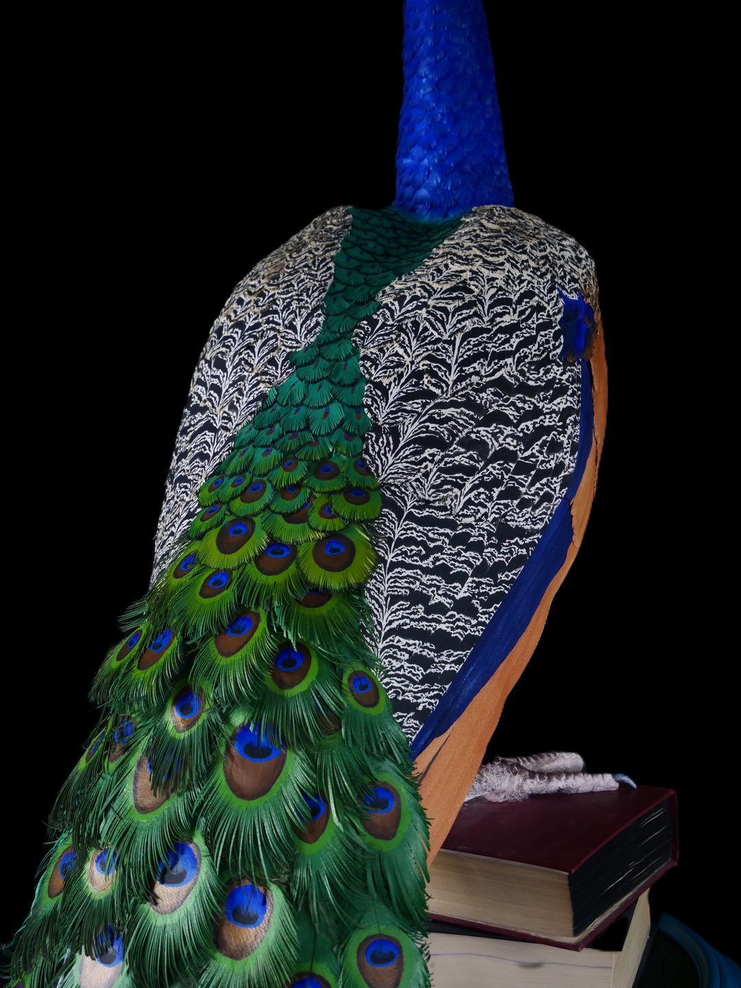 Arrogant Beauty by Jamie Lord – a life-size peacock sculpture meticulously crafted from hand-cut, hand-painted 260gsm pastel paper feathers. The peacock stands proudly atop stacked vintage books, its vivid green and blue plumage cascading nearly to the floor. Designed to symbolise pride, intelligence, and transformation, the sculpture features lifelike detail and rich texture, standing 1.75 metres tall and presented against a black backdrop.
