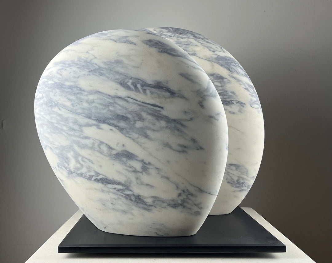 Jason Mulligan sculpture titled RISE, a one-off contemporary abstract artwork hand-carved from Portuguese marble. The sculpture features two large, rounded organic forms rising side by side, their surfaces polished smooth to reveal natural white marble with grey-blue veining. Presented on a black plinth, the piece embodies balance, unity, and quiet strength, blending modern minimalism with timeless stone carving traditions.