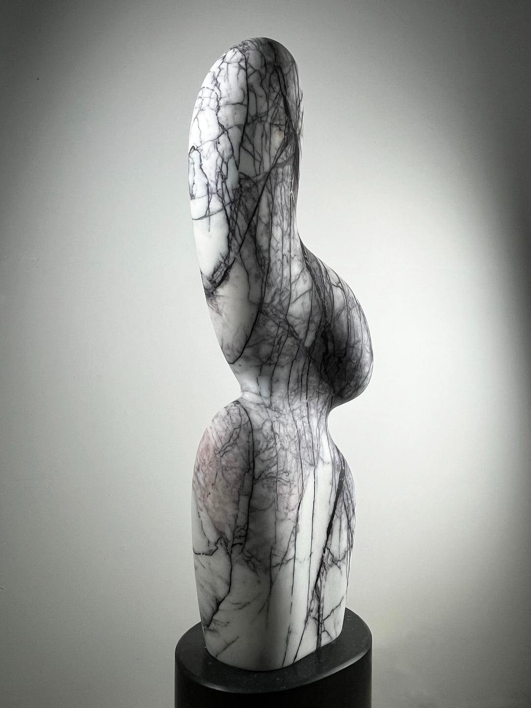 Jason Mulligan sculpture LILAC NOMAD, a one-off abstract form hand-carved from Turkish Lilac marble with a Kilkenny limestone base. Standing 1.3 metres tall, the sculpture rises with a flowing, organic silhouette that evokes movement and presence. The polished marble is veined with striking lilac, grey, and black patterns across a soft white ground, giving the work a layered depth and natural energy. Resting on dark Kilkenny limestone, this contemporary piece conveys elegance, strength, and timeless.
