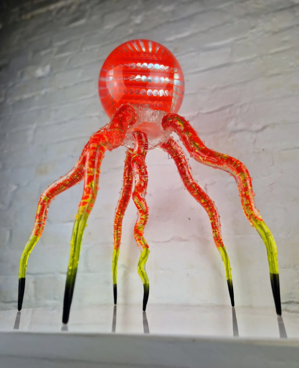 Futuristic jellyfish-like sculpture featuring a glowing spherical head and eight articulated legs. The top orb, made from neon orange acrylic and clear resin, contains layered geometric patterns that intensify under light. Below, segmented legs—formed from transparent rods in vibrant red, yellow, and lime green—extend to pointed black steel tips. The structure glows under illumination, casting vivid reflections and giving the impression of a living, biomechanical organism. Displayed against white brick wall