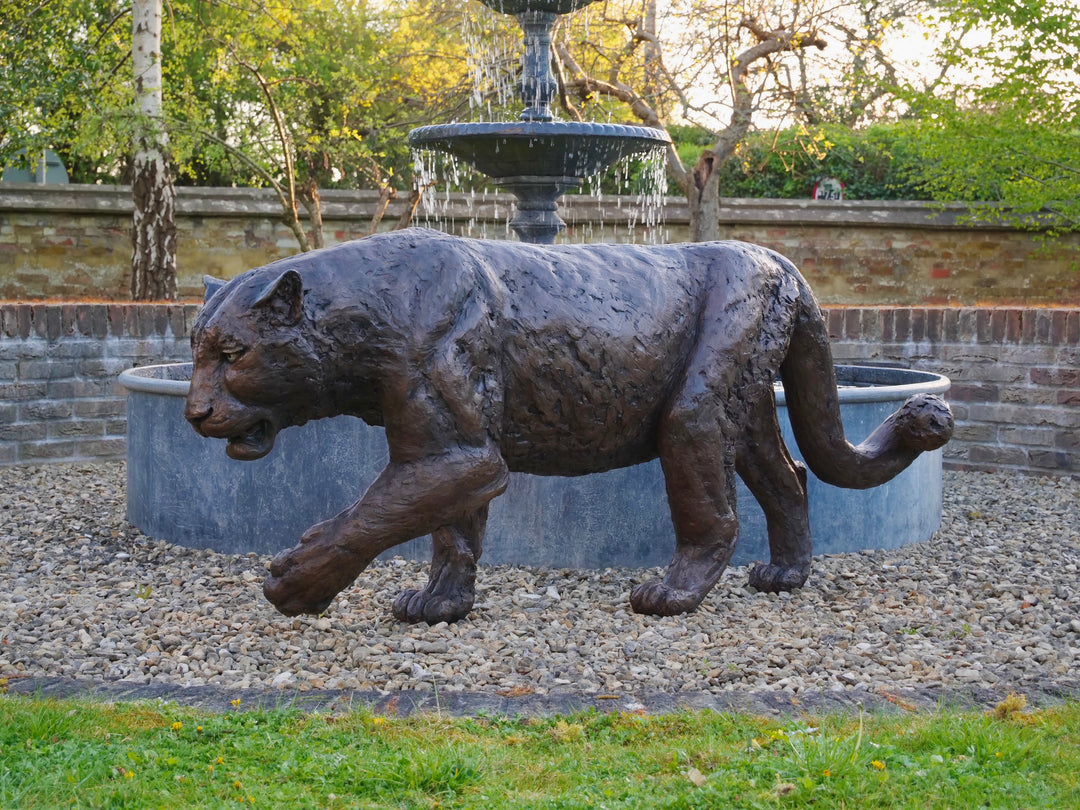 ‘Bloodline’ Amur leopard sculpture by Jamie Lord, life-size cold cast bronze of a leopard walking mid-stride with raised paw, textured dark bronze surface set outdoors on grass against green foliage