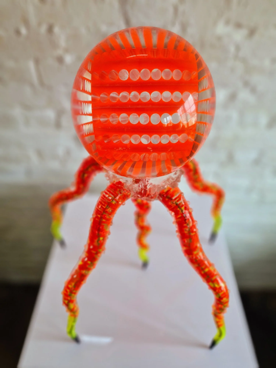 Futuristic jellyfish-like sculpture featuring a glowing spherical head and eight articulated legs. The top orb, made from neon orange acrylic and clear resin, contains layered geometric patterns that intensify under light. Below, segmented legs—formed from transparent rods in vibrant red, yellow, and lime green—extend to pointed black steel tips. The structure glows under illumination, casting vivid reflections and giving the impression of a living, biomechanical organism. Displayed against white brick wall