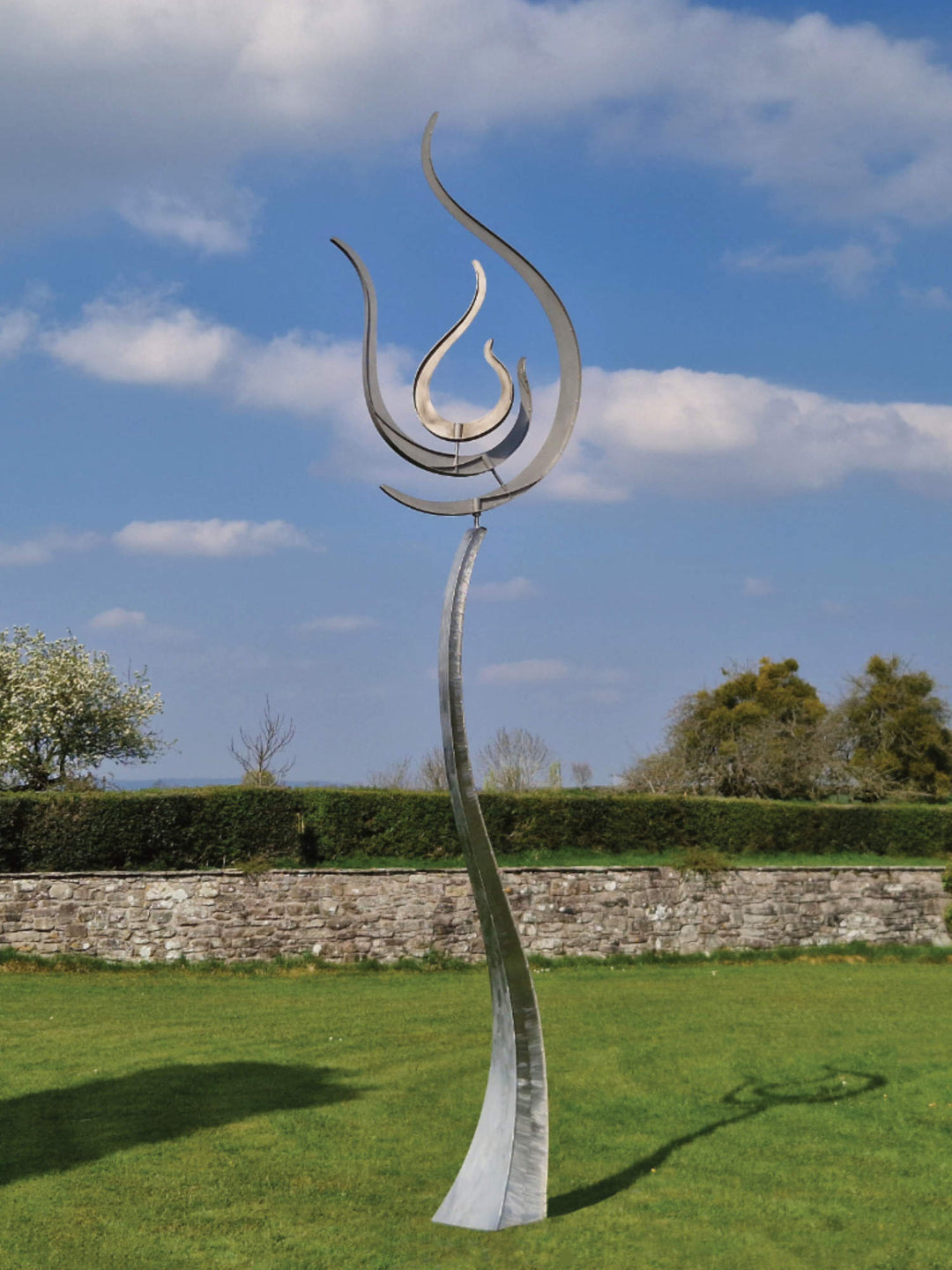 Tall stainless steel kinetic sculpture with fluid, flame-like curves, standing against a blue sky in a landscaped garden