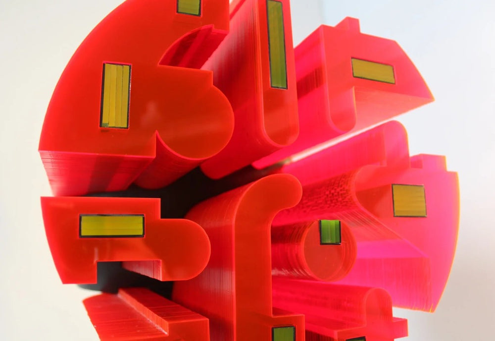 Futuristic sculptural form mounted on a black granite base, featuring a central spherical structure composed of layered neon red acrylic and matte black HIPS segments. The sculpture is precision-carved with bold geometric cut-outs and embedded reflective elements in green and yellow. Its striking contrast and luminous red glow give it the presence of an otherworldly artefact or machine, exuding personality and presence.