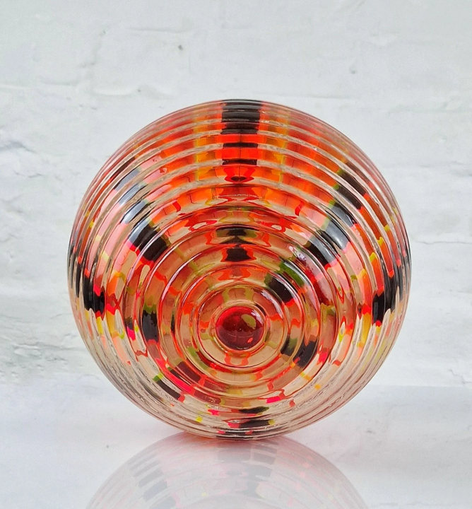Spherical resin and acrylic sculpture with concentric ridges and vibrant internal colour patterns. The layered composition features fluorescent red, yellow, black and orange acrylic bands encapsulated in clear resin, creating an optical effect that shifts with light and angle. Displayed on a white plinth against a white brick wall, the piece radiates an energetic, cosmic presence—evoking portals, celestial forms, or futuristic artefacts.