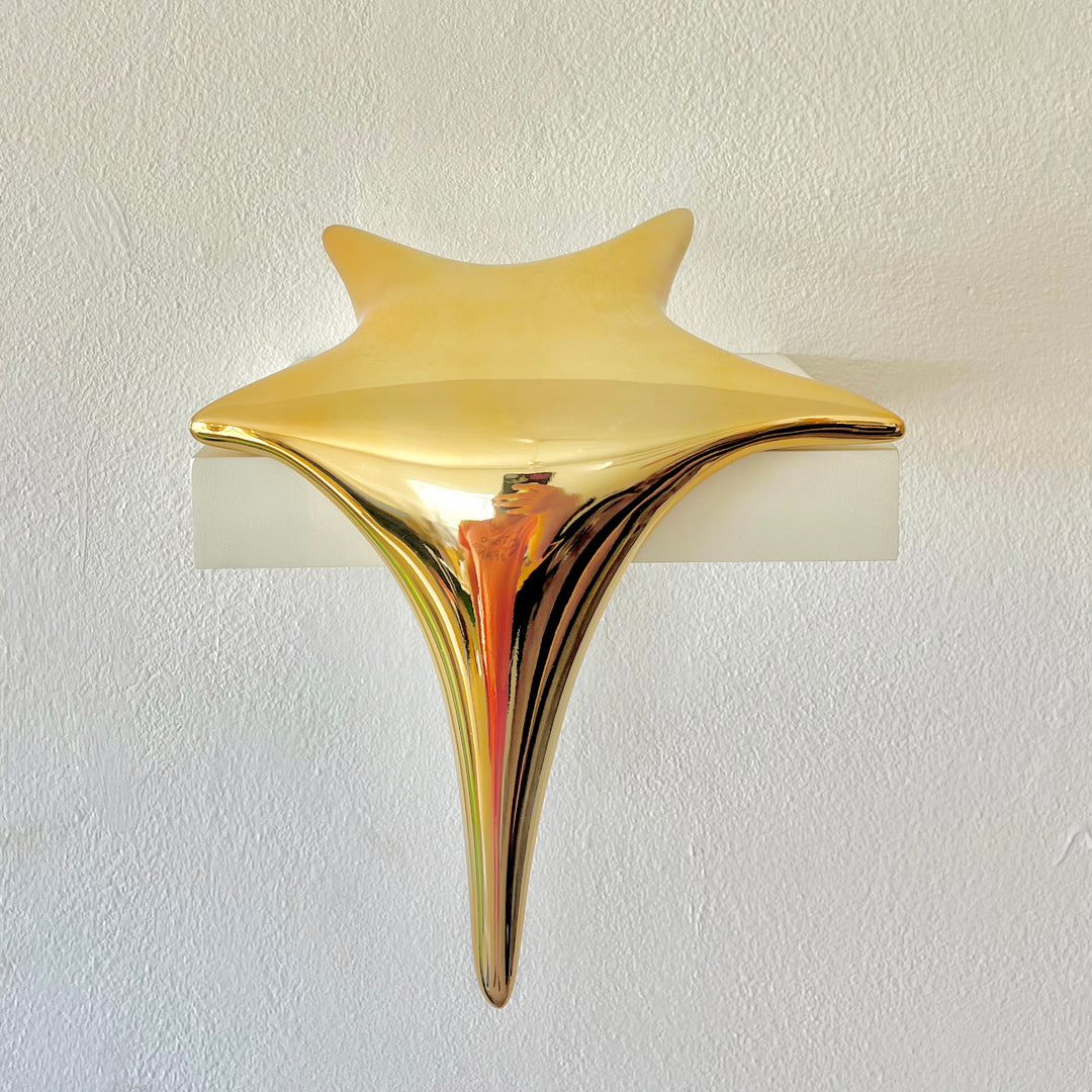 Polished gold-toned stainless steel sculpture in the form of a stylised shooting star, with a fluid, elongated spike melting over the edge of a white plinth or shelf.