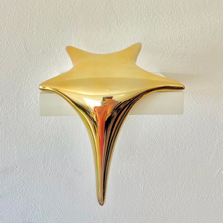 Polished gold-toned stainless steel sculpture in the form of a stylised shooting star, with a fluid, elongated spike melting over the edge of a white plinth or shelf.
