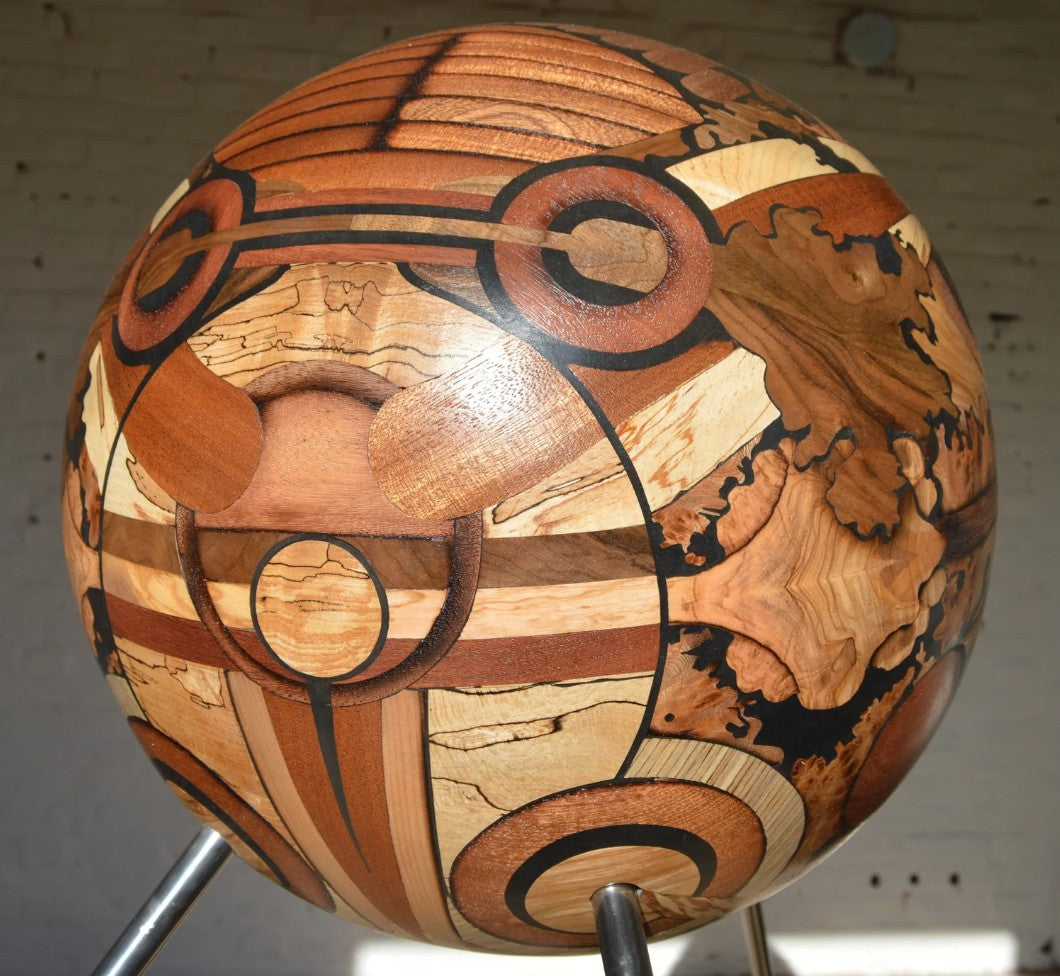 Spherical mixed wood sculpture by Richard Slee titled “RA,” featuring intricate marquetry in warm wood tones with milliput detailing. Raised on three slender steel legs, the large-scale work casts dynamic shadows and evokes planetary or celestial form with layered textures and radial balance.