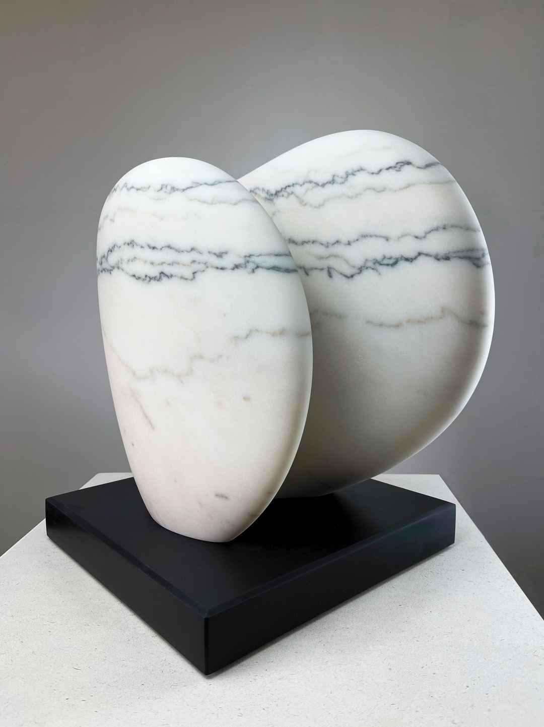 Jason Mulligan sculpture titled ECLIPSE, a one-off hand-carved abstract artwork in Portuguese marble. The piece features two interlocking, rounded organic forms with a smooth polished surface, revealing the natural white marble streaked with striking grey linear veining. Resting on a dark plinth, the sculpture embodies harmony, duality, and celestial balance, evoking the timeless elegance of stone carving through a contemporary minimalist lens.