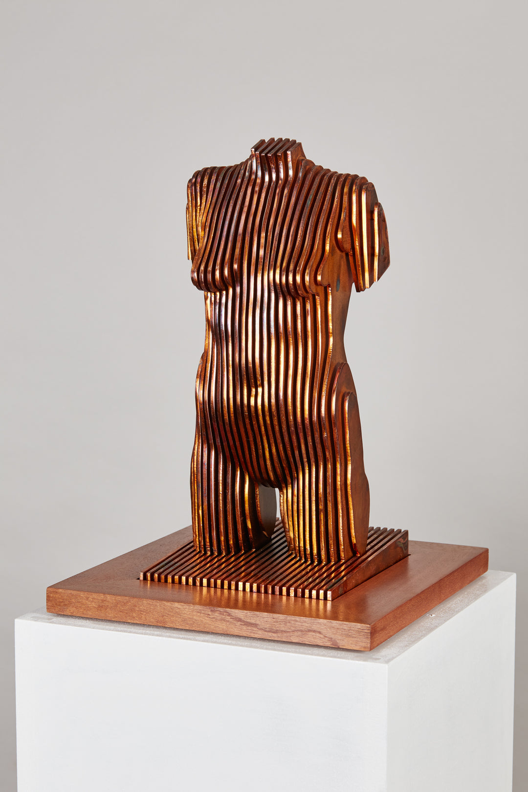 Collaborative sculpture by Tom Hiscox and Fred Baier featuring a stylised female torso formed from layered corten steel and copper plate, mounted on a geometric American walnut and oak base, shown in a light-filled interior setting.