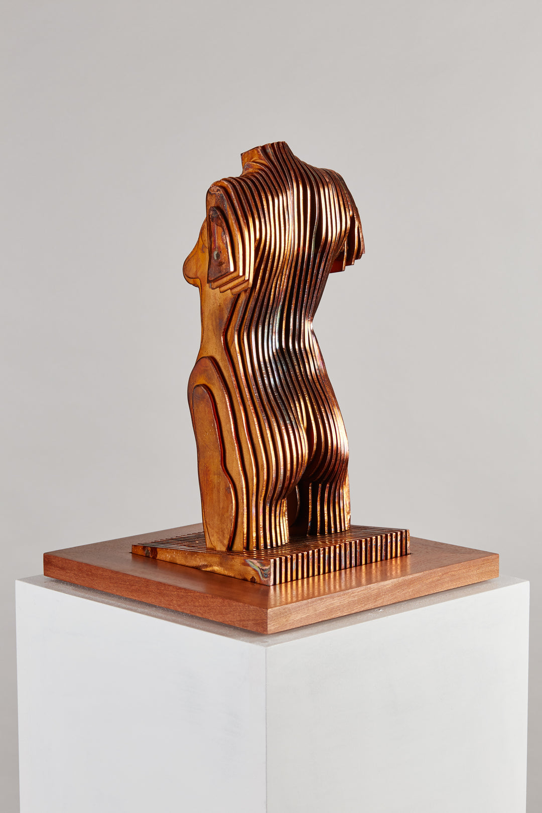 Collaborative sculpture by Tom Hiscox and Fred Baier featuring a stylised female torso formed from layered corten steel and copper plate, mounted on a geometric American walnut and oak base, shown in a light-filled interior setting.