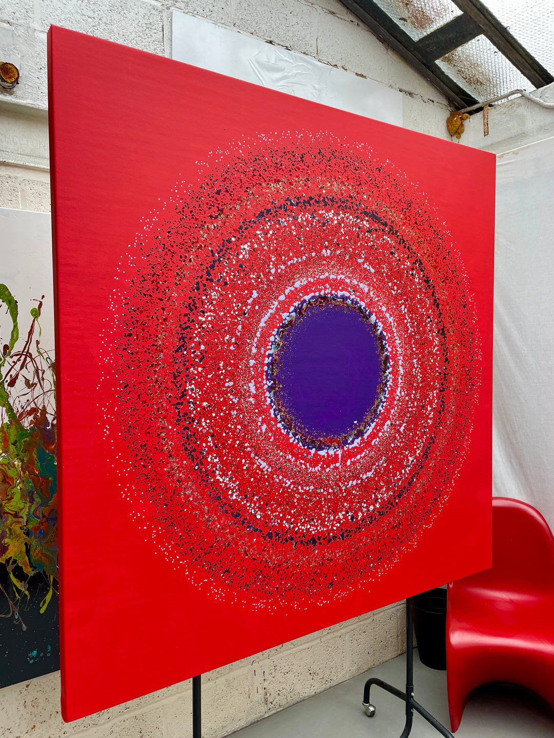 Large abstract painting “Ellipsis” by Swarez — bold square composition with a deep violet central circle surrounded by concentric rings of paint droplets and splashes on a vivid matte red background. Energetic, dynamic halo effect created by spin technique.