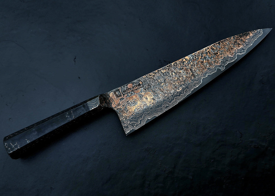 Close-up of a hand-forged Cu-Ni-Mascus Gyuto chef’s knife by Leszek Sikon, featuring an intricate 8120-layer Damascus pattern combining copper, nickel, brass, and high-carbon steels. The blade has a molten, marbled texture with visible copper sheen and an S-profile for food release, paired with a custom jeans micarta handle with embedded copper and steel mesh.