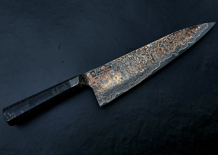 Close-up of a hand-forged Cu-Ni-Mascus Gyuto chef’s knife by Leszek Sikon, featuring an intricate 8120-layer Damascus pattern combining copper, nickel, brass, and high-carbon steels. The blade has a molten, marbled texture with visible copper sheen and an S-profile for food release, paired with a custom jeans micarta handle with embedded copper and steel mesh.