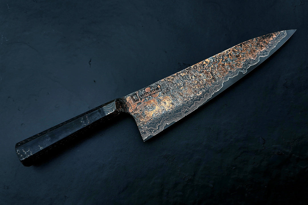 Close-up of a hand-forged Cu-Ni-Mascus Gyuto chef’s knife by Leszek Sikon, featuring an intricate 8120-layer Damascus pattern combining copper, nickel, brass, and high-carbon steels. The blade has a molten, marbled texture with visible copper sheen and an S-profile for food release, paired with a custom jeans micarta handle with embedded copper and steel mesh.