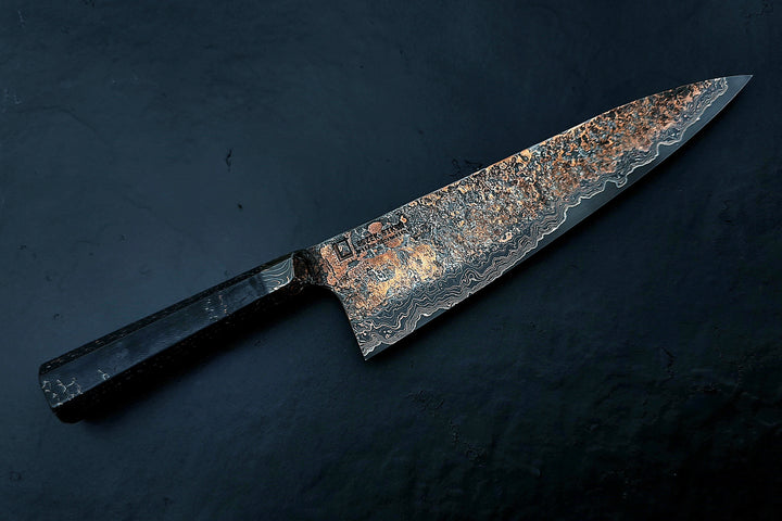 Close-up of a hand-forged Cu-Ni-Mascus Gyuto chef’s knife by Leszek Sikon, featuring an intricate 8120-layer Damascus pattern combining copper, nickel, brass, and high-carbon steels. The blade has a molten, marbled texture with visible copper sheen and an S-profile for food release, paired with a custom jeans micarta handle with embedded copper and steel mesh.