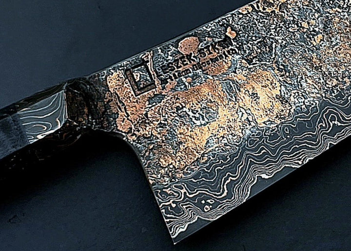 Close-up of a hand-forged Cu-Ni-Mascus Gyuto chef’s knife by Leszek Sikon, featuring an intricate 8120-layer Damascus pattern combining copper, nickel, brass, and high-carbon steels. The blade has a molten, marbled texture with visible copper sheen and an S-profile for food release, paired with a custom jeans micarta handle with embedded copper and steel mesh.