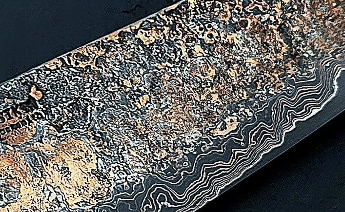 Close-up of a hand-forged Cu-Ni-Mascus Gyuto chef’s knife by Leszek Sikon, featuring an intricate 8120-layer Damascus pattern combining copper, nickel, brass, and high-carbon steels. The blade has a molten, marbled texture with visible copper sheen and an S-profile for food release, paired with a custom jeans micarta handle with embedded copper and steel mesh.