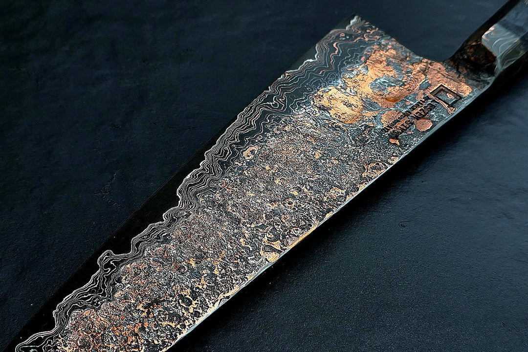 Close-up of a hand-forged Cu-Ni-Mascus Gyuto chef’s knife by Leszek Sikon, featuring an intricate 8120-layer Damascus pattern combining copper, nickel, brass, and high-carbon steels. The blade has a molten, marbled texture with visible copper sheen and an S-profile for food release, paired with a custom jeans micarta handle with embedded copper and steel mesh.