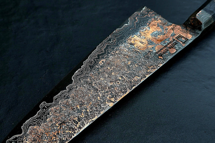 Close-up of a hand-forged Cu-Ni-Mascus Gyuto chef’s knife by Leszek Sikon, featuring an intricate 8120-layer Damascus pattern combining copper, nickel, brass, and high-carbon steels. The blade has a molten, marbled texture with visible copper sheen and an S-profile for food release, paired with a custom jeans micarta handle with embedded copper and steel mesh.
