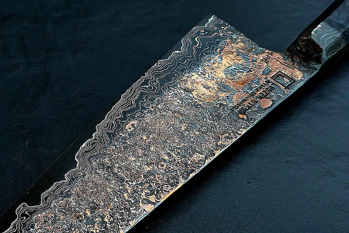 Close-up of a hand-forged Cu-Ni-Mascus Gyuto chef’s knife by Leszek Sikon, featuring an intricate 8120-layer Damascus pattern combining copper, nickel, brass, and high-carbon steels. The blade has a molten, marbled texture with visible copper sheen and an S-profile for food release, paired with a custom jeans micarta handle with embedded copper and steel mesh.
