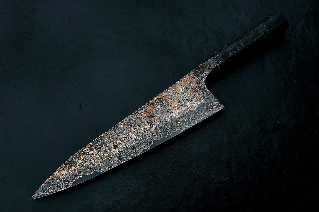 Close-up of a hand-forged Cu-Ni-Mascus Gyuto chef’s knife by Leszek Sikon, featuring an intricate 8120-layer Damascus pattern combining copper, nickel, brass, and high-carbon steels. The blade has a molten, marbled texture with visible copper sheen and an S-profile for food release, paired with a custom jeans micarta handle with embedded copper and steel mesh.