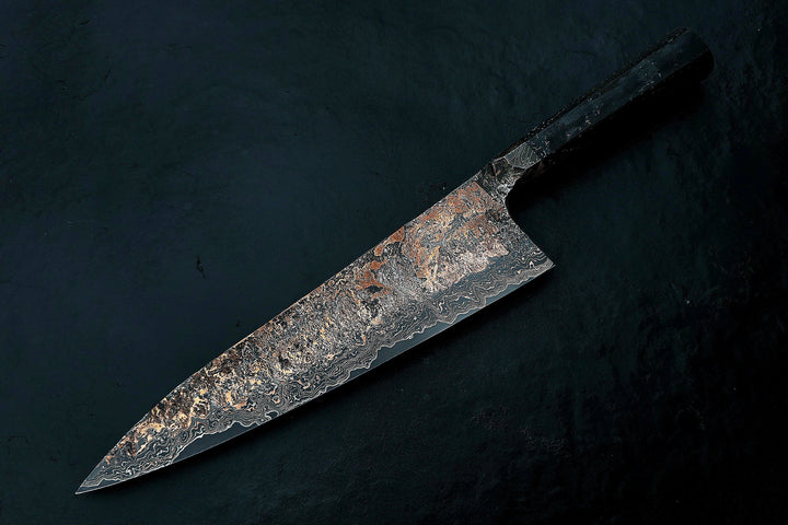 Close-up of a hand-forged Cu-Ni-Mascus Gyuto chef’s knife by Leszek Sikon, featuring an intricate 8120-layer Damascus pattern combining copper, nickel, brass, and high-carbon steels. The blade has a molten, marbled texture with visible copper sheen and an S-profile for food release, paired with a custom jeans micarta handle with embedded copper and steel mesh.