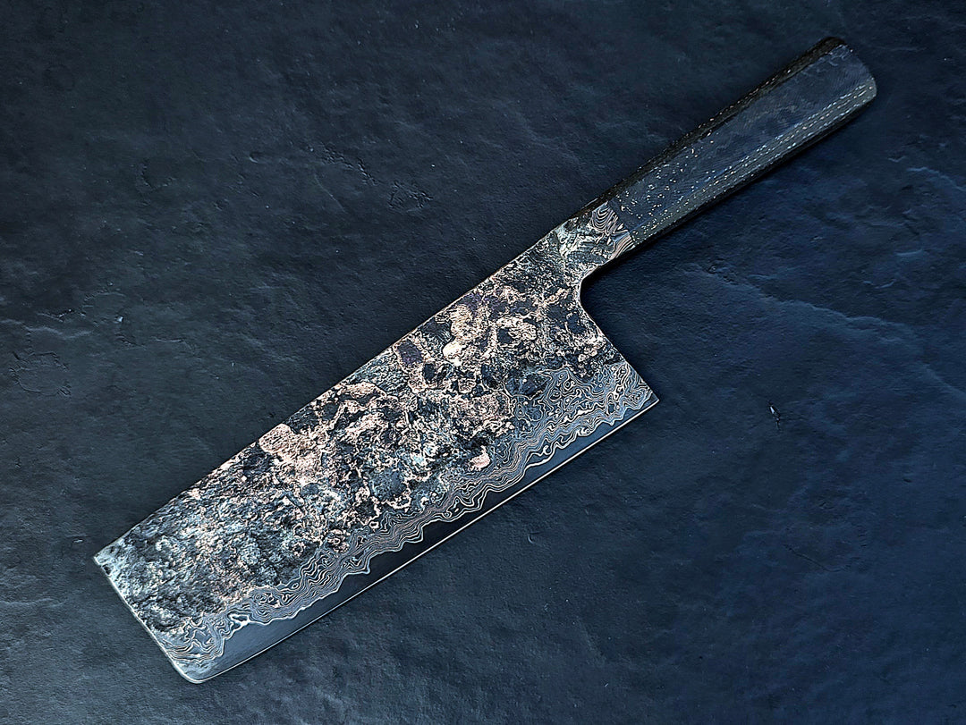 One-of-a-kind Cu-Ni-Mascus Nakiri knife by Leszek Sikon, showcasing 8120 layers of Damascus forged from Spicy White steel, O1 tool steel, copper, nickel, and brass. The blade features a rich, geological pattern with molten textures and wave-like lines, hand-forged over 100 hours with an S-profile for food release and finished with a custom jeans micarta handle embedded with copper and steel mesh.